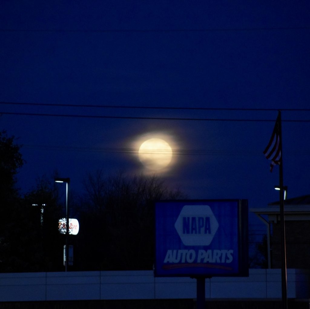 A full, large golden mood in a dark blue sky among business signs and flagpole with an American flag