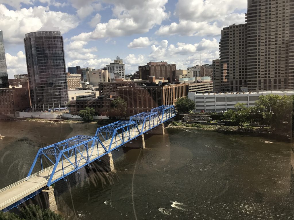 View of a blue-girder pedestrian bridge over the Grand River with a cityscape on the other side of the river.
