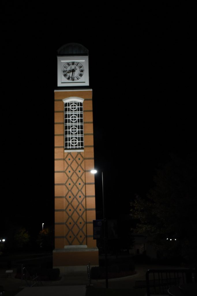 A terra cotta colored brick clock tower with dark green and white detailing and a white face at night.