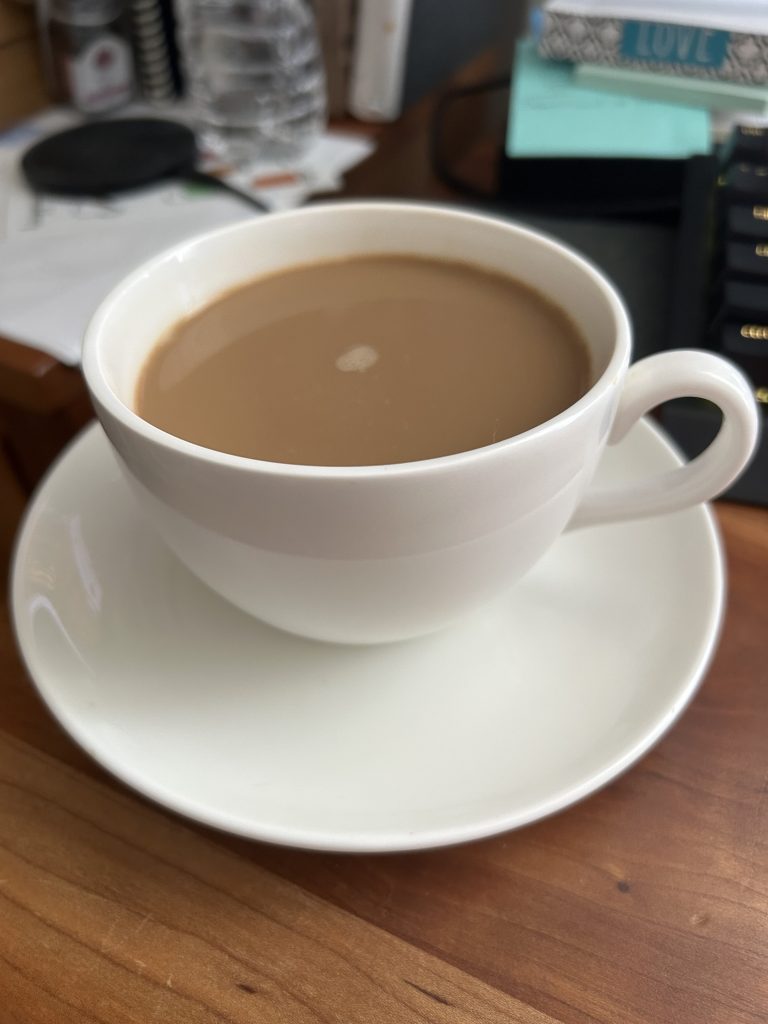 Close shot of a cream-colored cup and saucer filled with coffee with cream on a messy, modern desk.