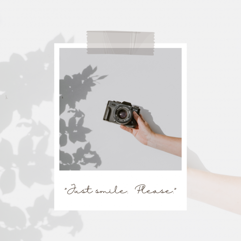 A hand holding a camera inside of a polaroid frame. Words in black read "Just smile. Please."