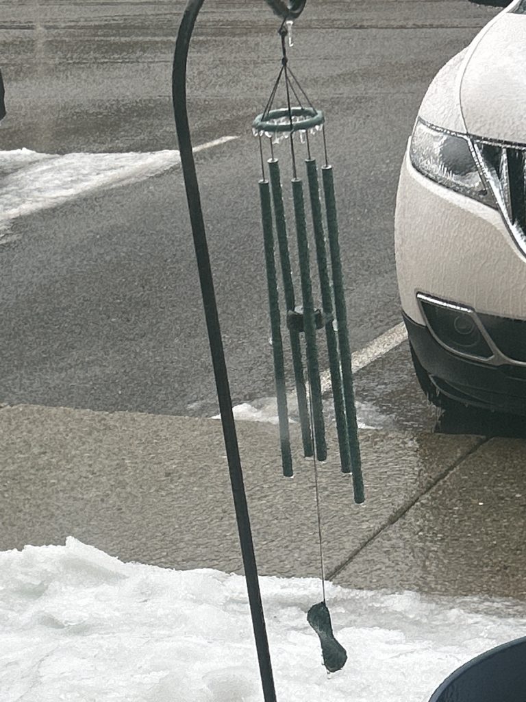 A set of green metal wind chimes on a shepherd’s crook coated in ice.