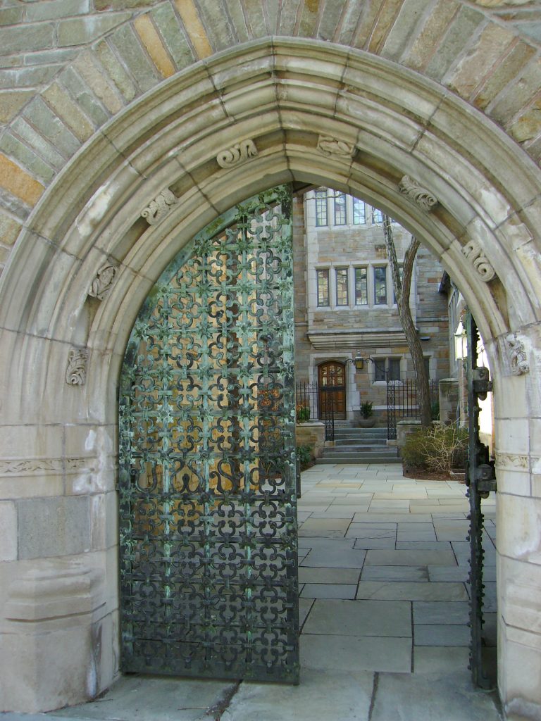 An old building in light stone seen through an archway with an oxidized grate that stands half open.