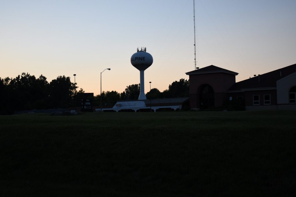 Clear-sky sunset over a building and parking lot and trees with a radio tower and water tower.