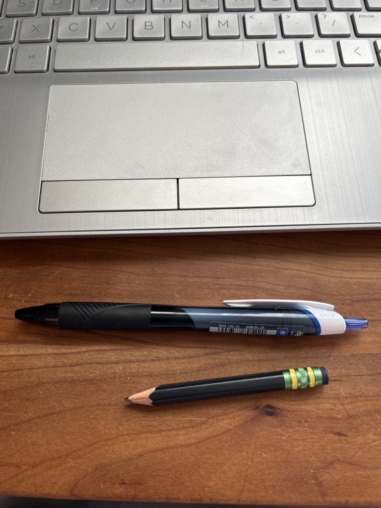 A close up of a laptop touchpad, a pen, and a black pencil that’s been sharpened down to a tiny nub on a wooden desk.