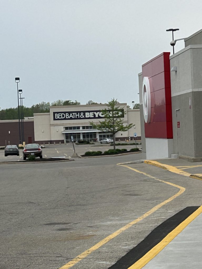 An empty Bed Bath and Beyond storefront seen across a parking lot. Target sign in the foreground on an angle.