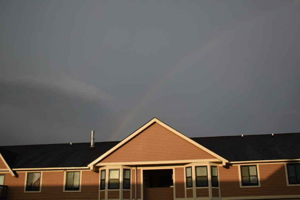 Rainbow against a dark sky over an apartment building front lit by the sun.