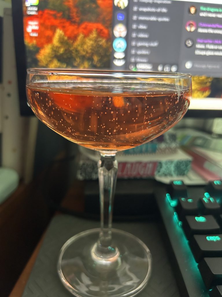 A coupe glass filled with pink sparkling wine in front of a computer screen.